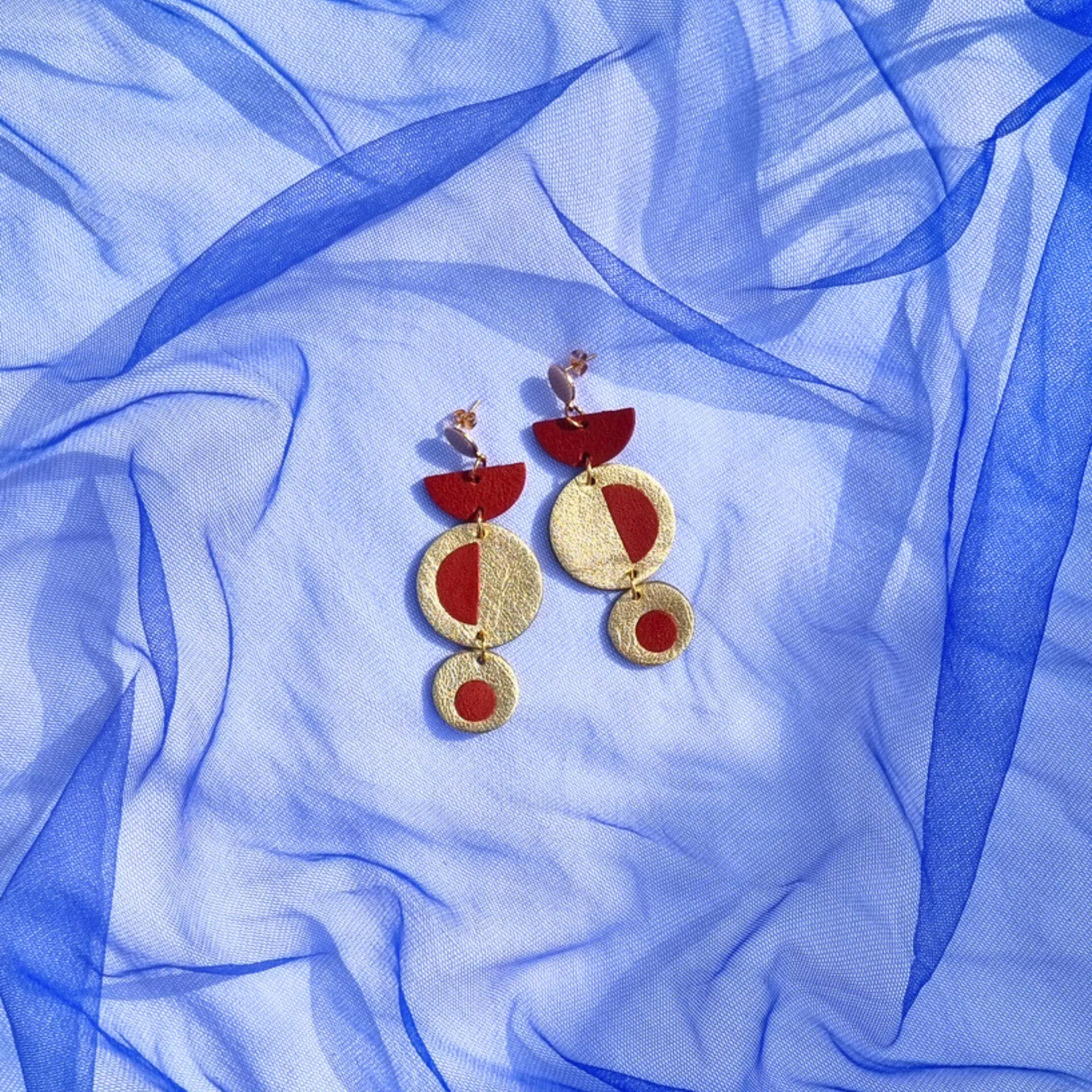 Handcrafted geometric dangly earrings with brick red inlayed in metallic gold leather, on a royal blue mesh fabric background.