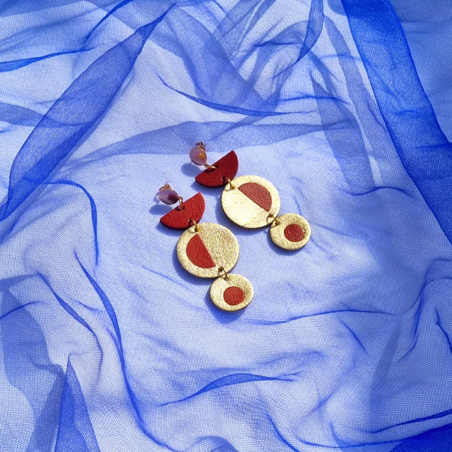 Handcrafted geometric dangly earrings with brick red inlayed in metallic gold leather, on a royal blue mesh fabric background.