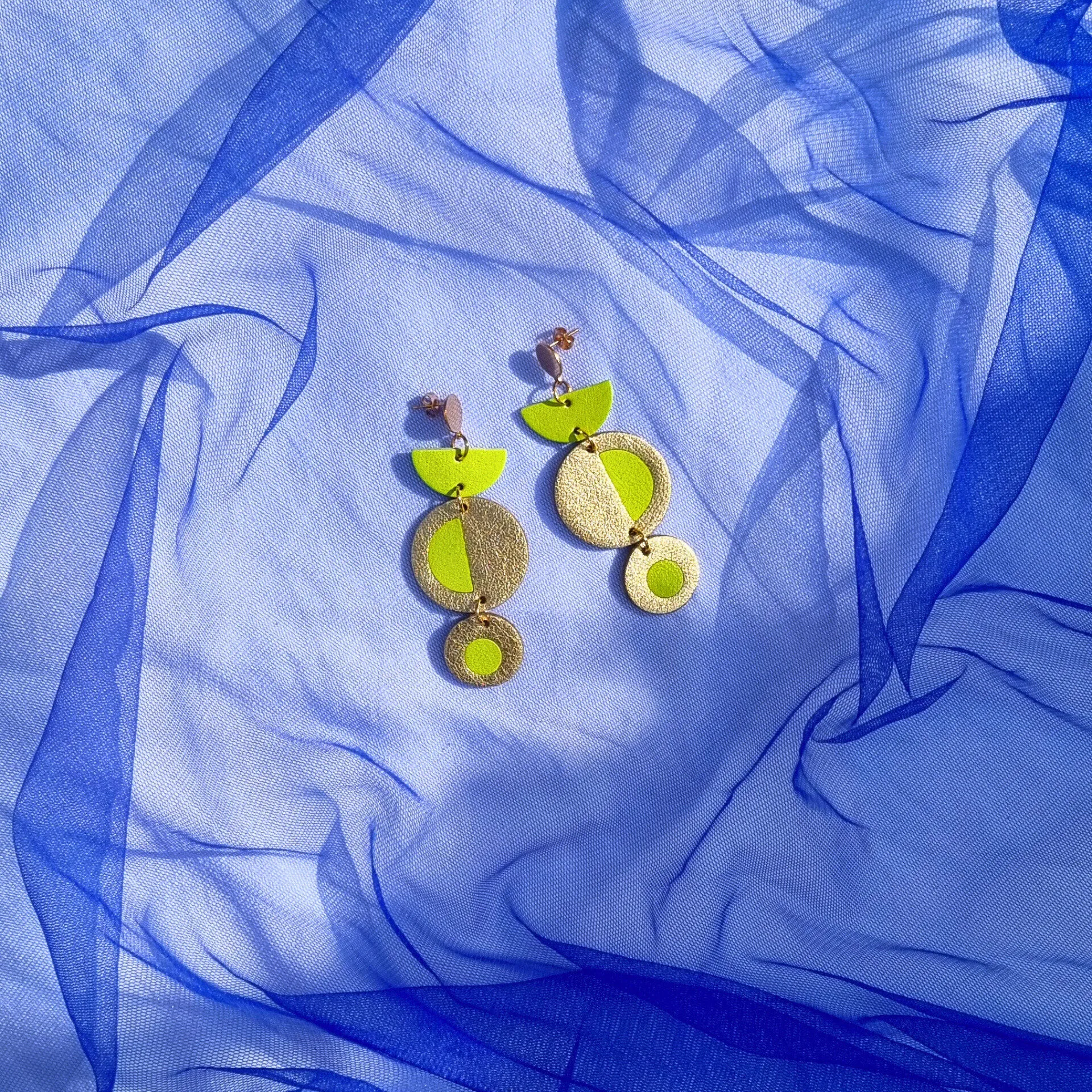 Geometric acid yellow and gold leather dangly earrings featuring a circular inlay design, resting on a draped bright blue tulle fabric background.