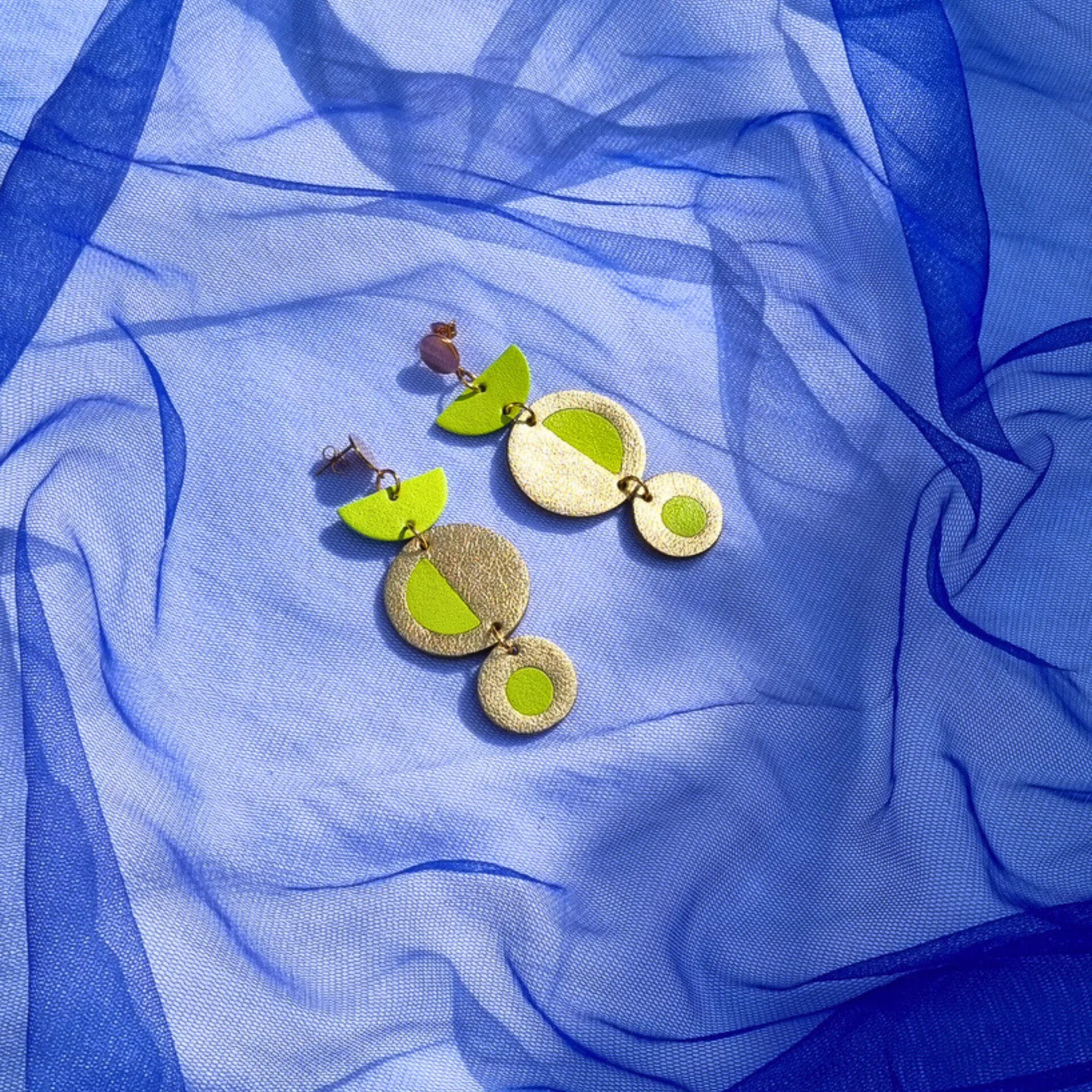 Geometric acid yellow and gold leather dangly earrings featuring a circular inlay design, resting on a draped bright blue tulle fabric background.