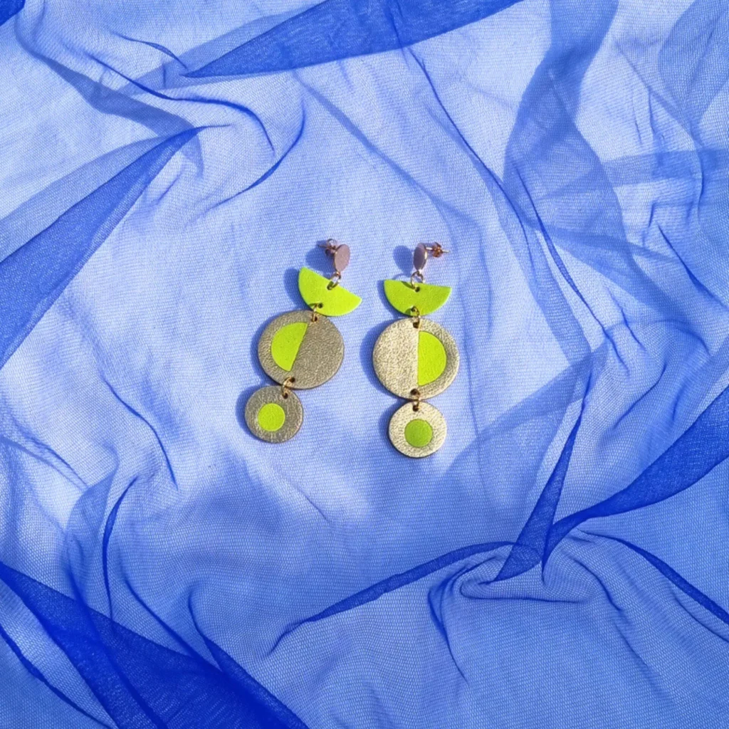 Geometric acid yellow and gold leather dangly earrings featuring a circular inlay design, resting on a draped bright blue tulle fabric background.