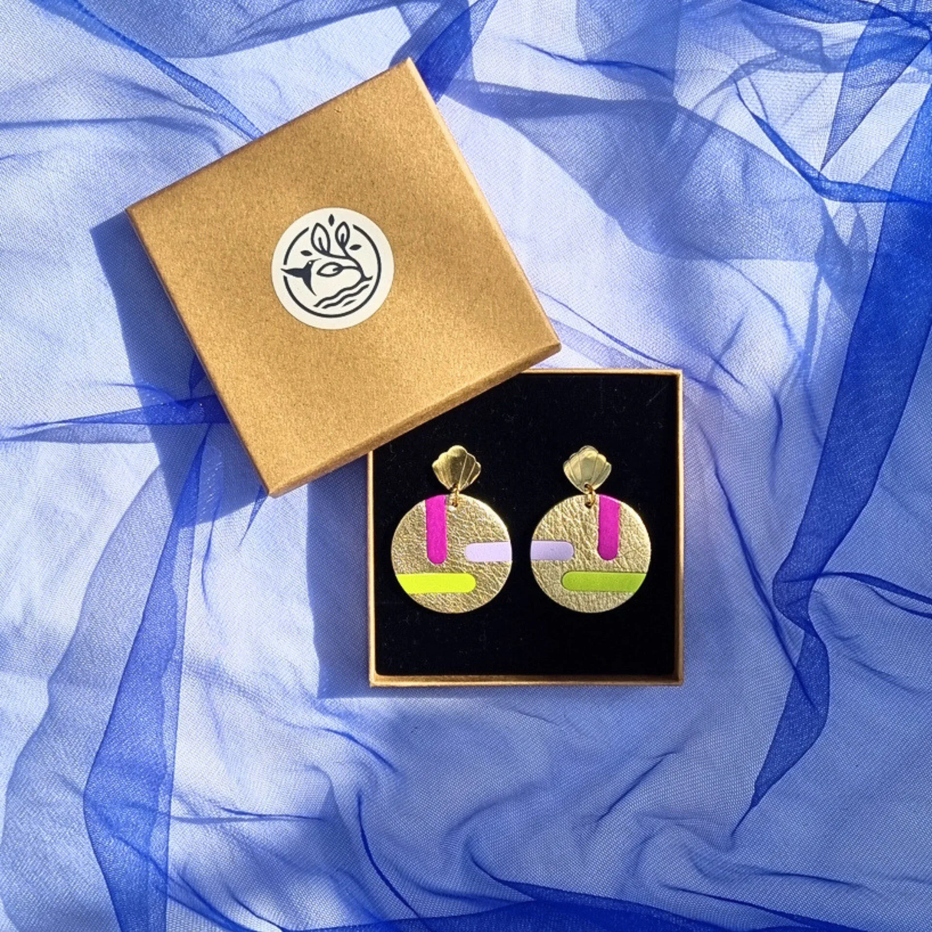 Circular gold leather disc earrings with abstract magenta, lilac and acid yellow inlays, displayed in a kraft gift box on a draped blue mesh fabric. The studs feature a gold shell shape.