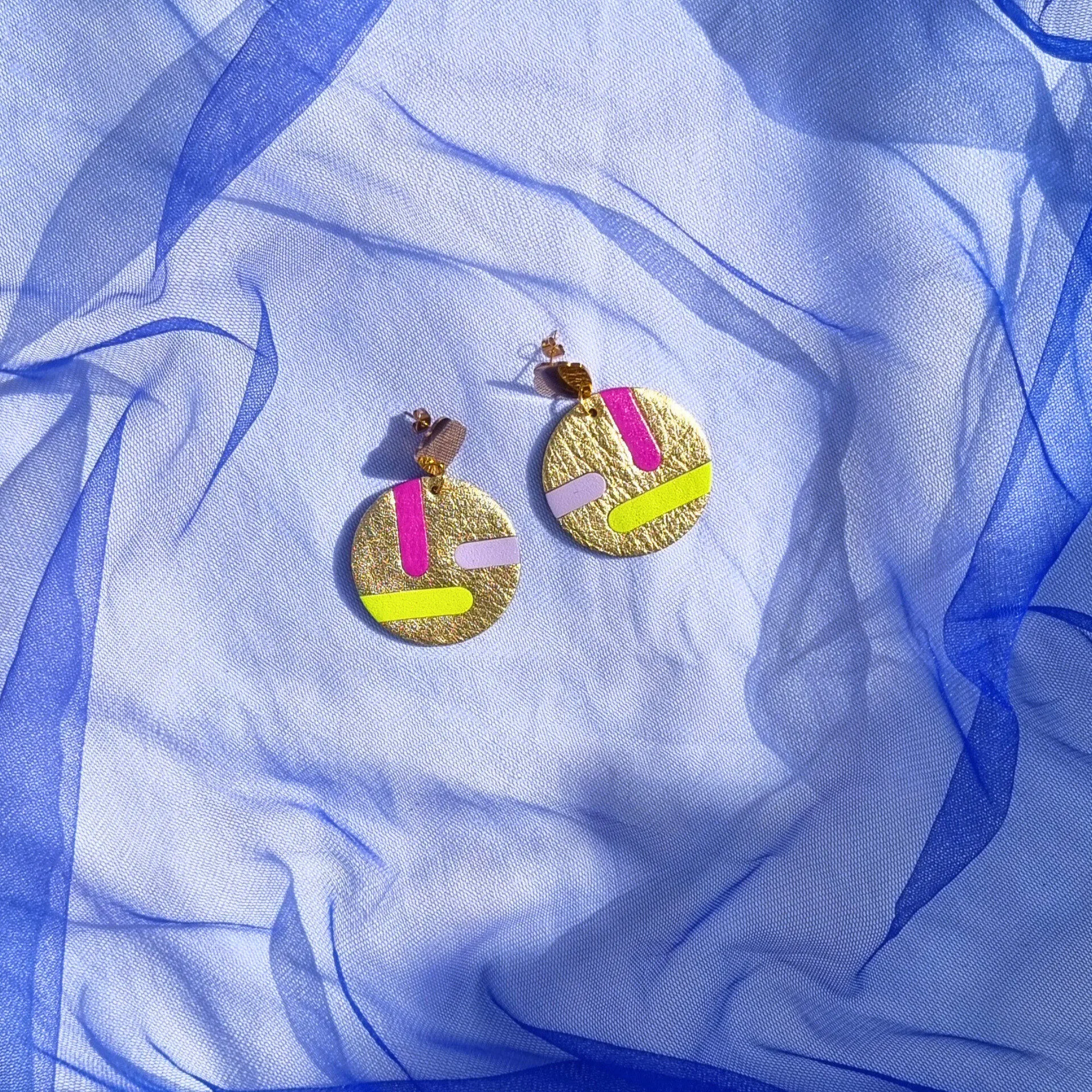 Circular gold leather disc earrings with abstract magenta, lilac and acid yellow inlays, displayed on a draped blue mesh fabric. The studs feature a gold shell shape.