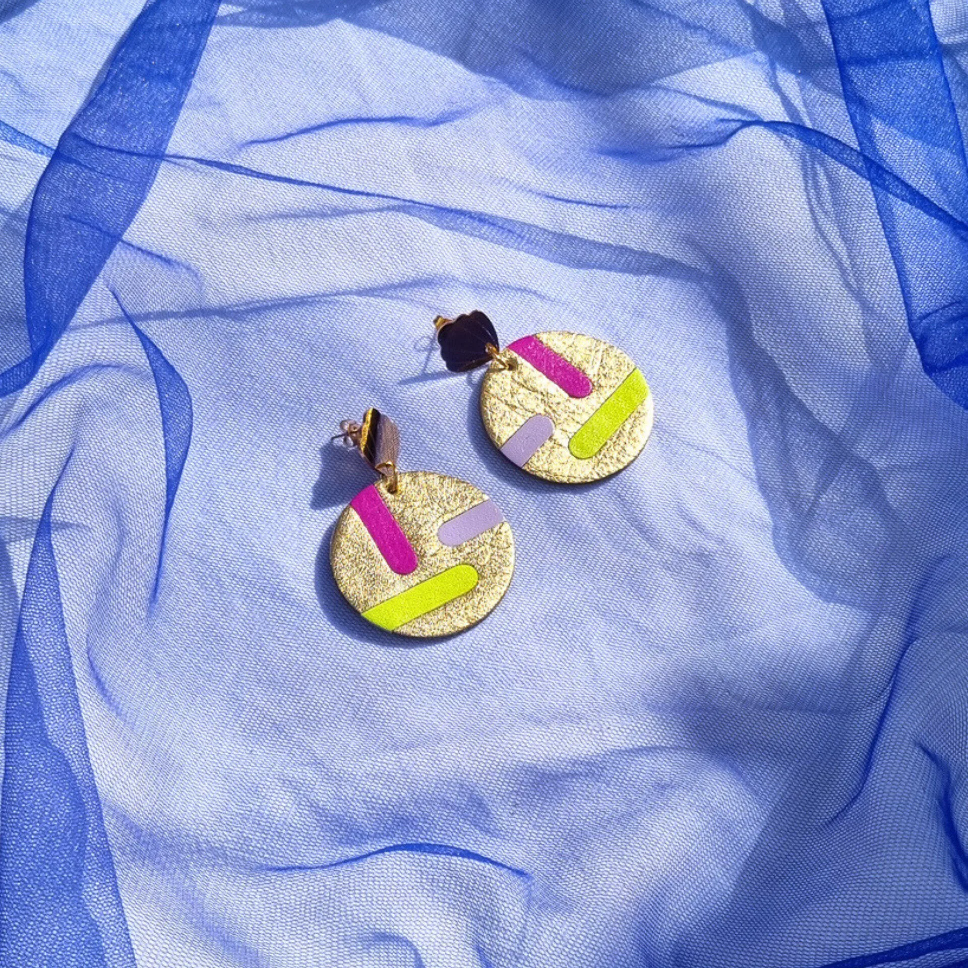 Circular gold leather disc earrings with abstract magenta, lilac and acid yellow inlays, displayed on a draped blue mesh fabric. The studs feature a gold shell shape.
