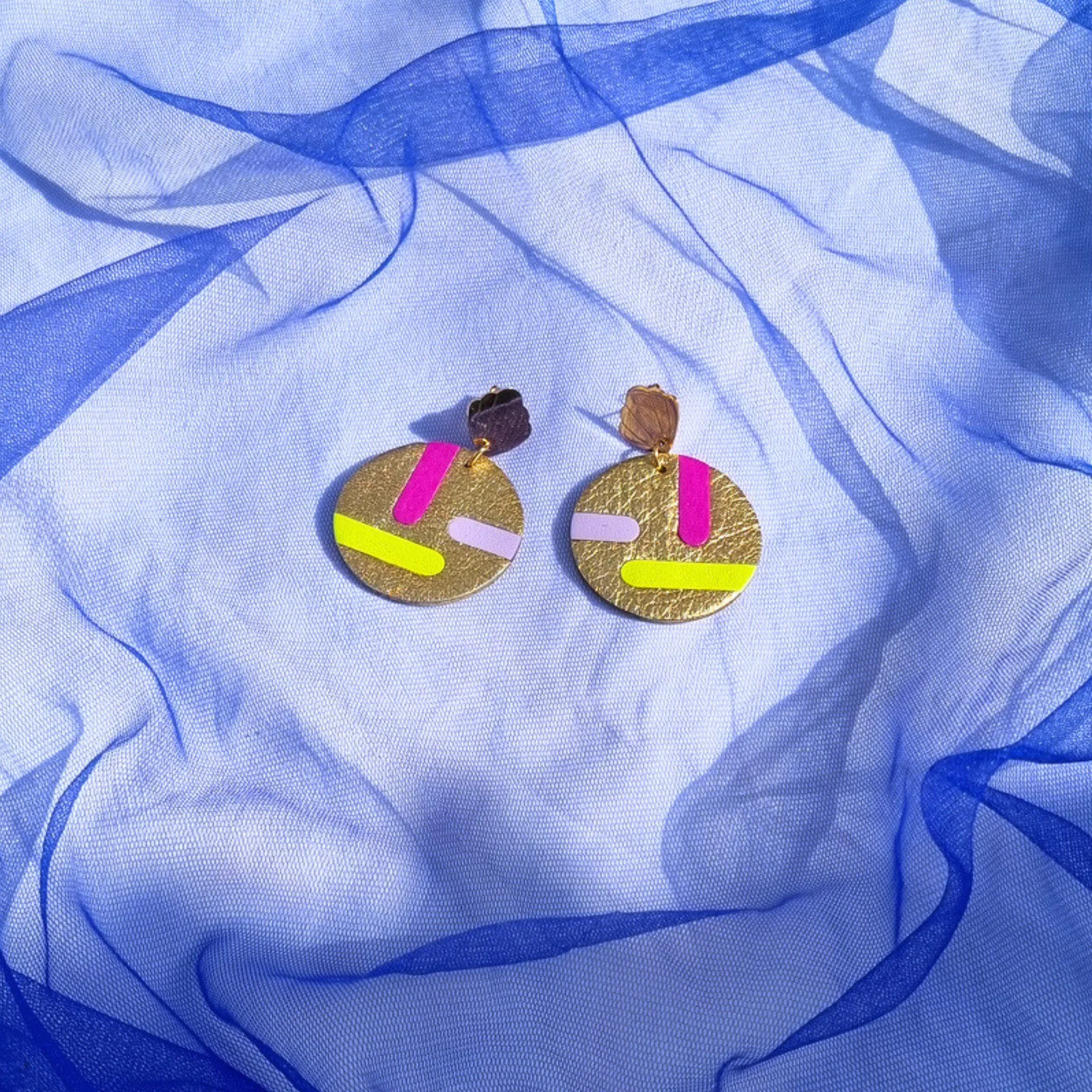 Circular gold leather disc earrings with abstract magenta, lilac and acid yellow inlays, displayed on a draped blue mesh fabric. The studs feature a gold shell shape.