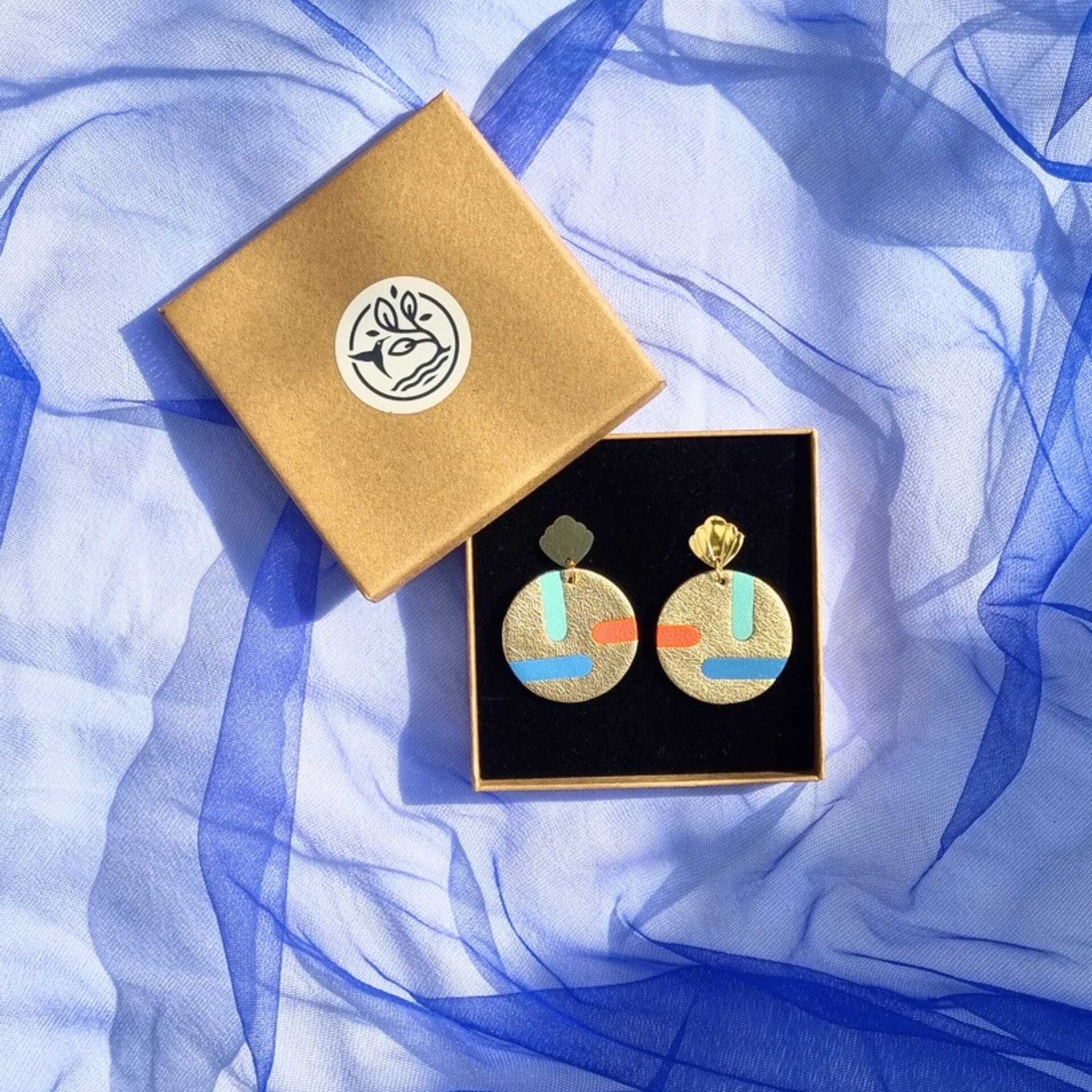 Circular gold leather disc earrings with abstract teal, blue, and orange inlays, displayed in a kraft gift box on a draped blue mesh fabric. The studs feature a gold shell shape.