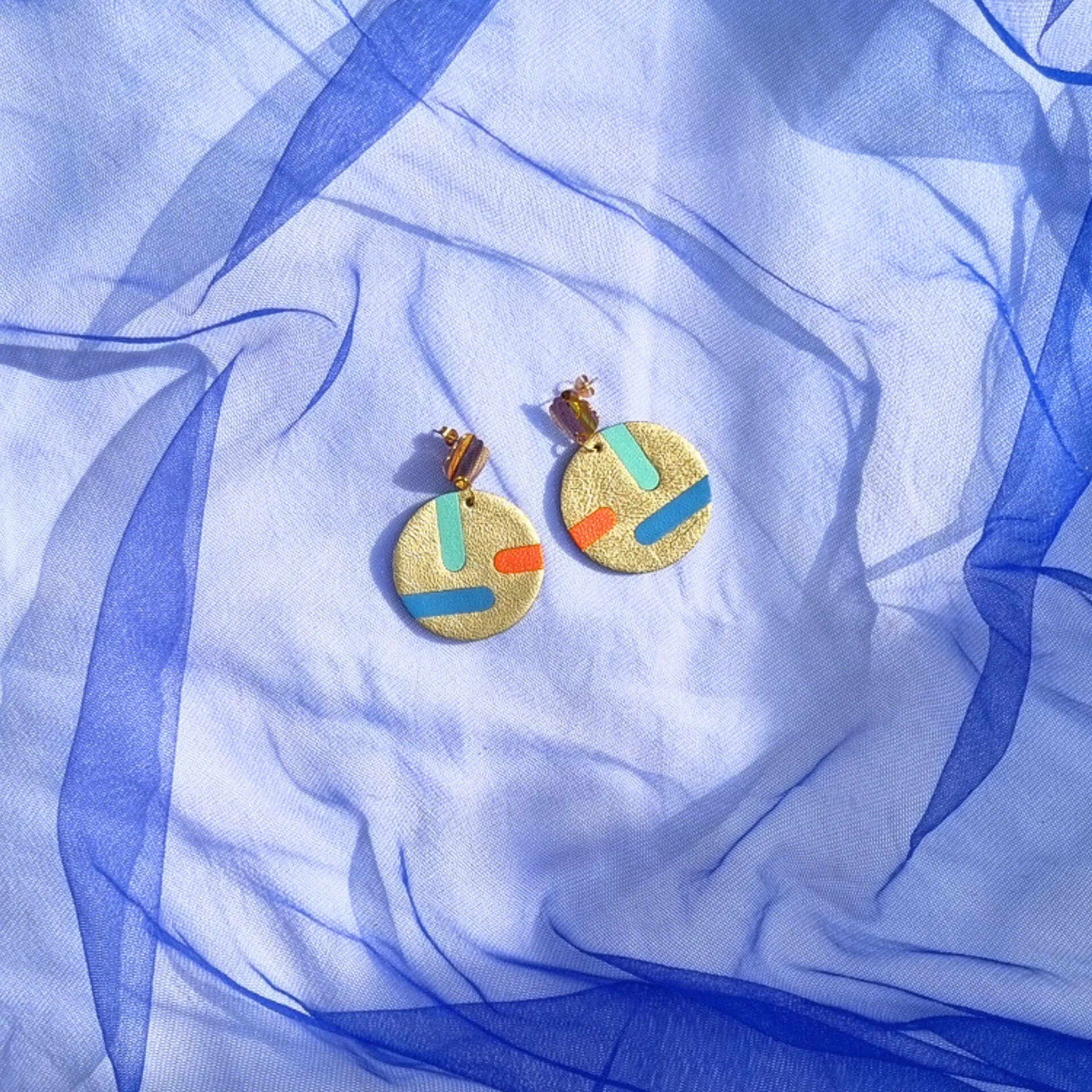 Circular gold leather disc earrings with abstract teal, blue, and orange inlays, displayed on a draped blue mesh fabric. The studs feature a gold shell shape.