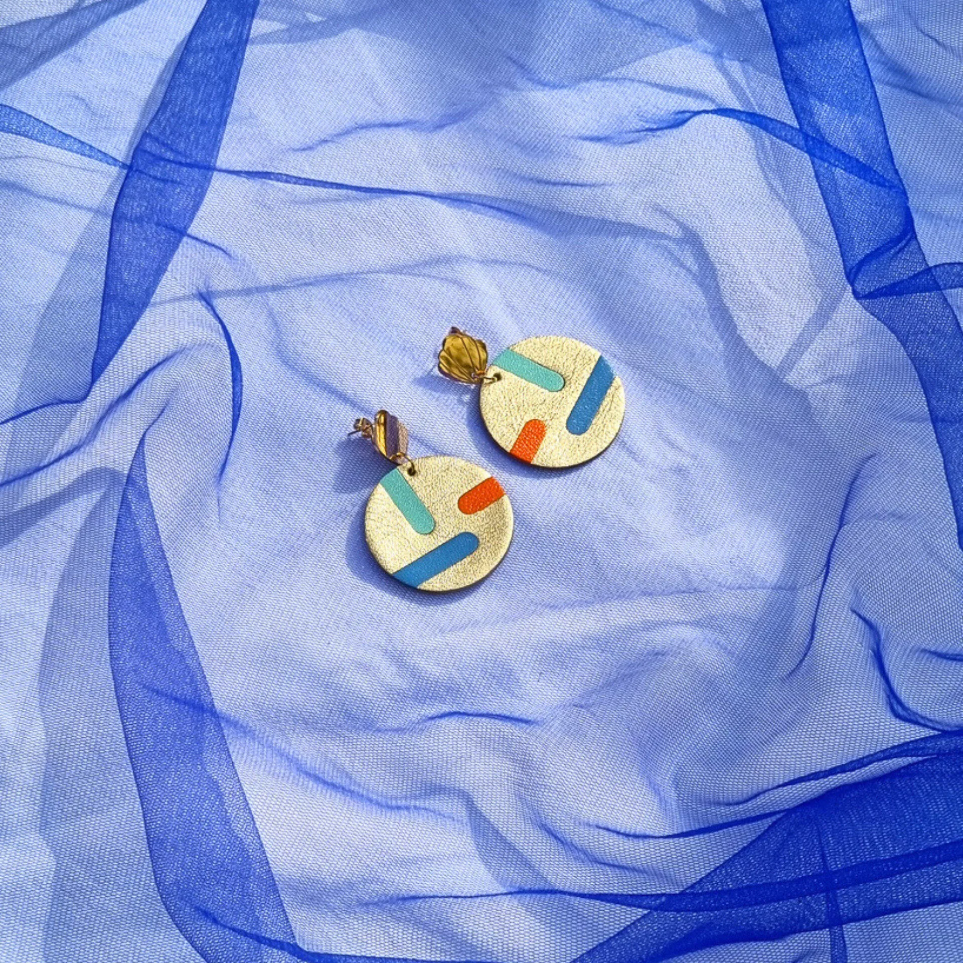 Circular gold leather disc earrings with abstract teal, blue, and orange inlays, displayed on a draped blue mesh fabric. The studs feature a gold shell shape.