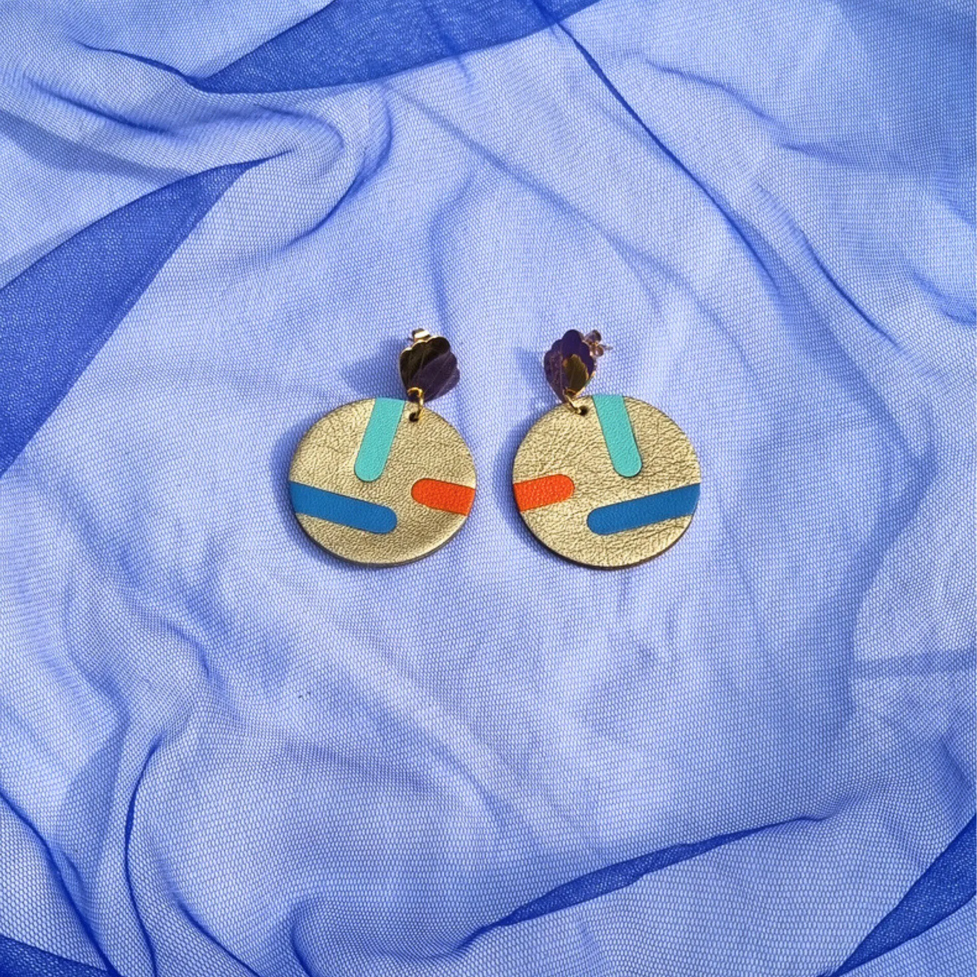 Circular gold leather disc earrings with abstract teal, blue, and orange inlays, displayed on a draped blue mesh fabric. The studs feature a gold shell shape.