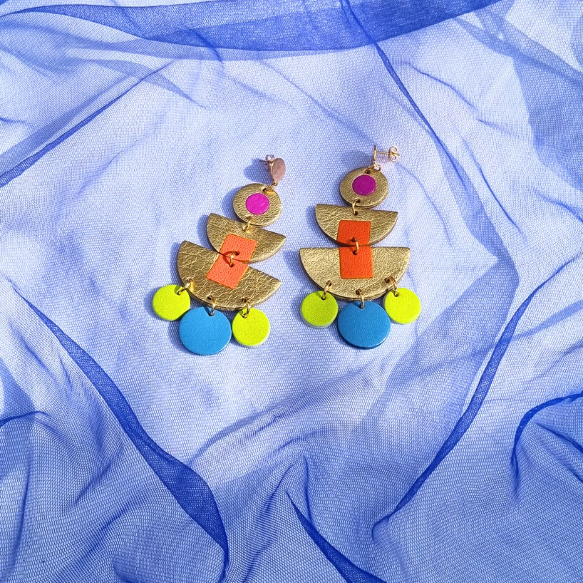 Geometric "Goddess" statement earrings on blue mesh fabric. The tiered gold leather design features vibrant magenta, orange, green, and blue inlays.