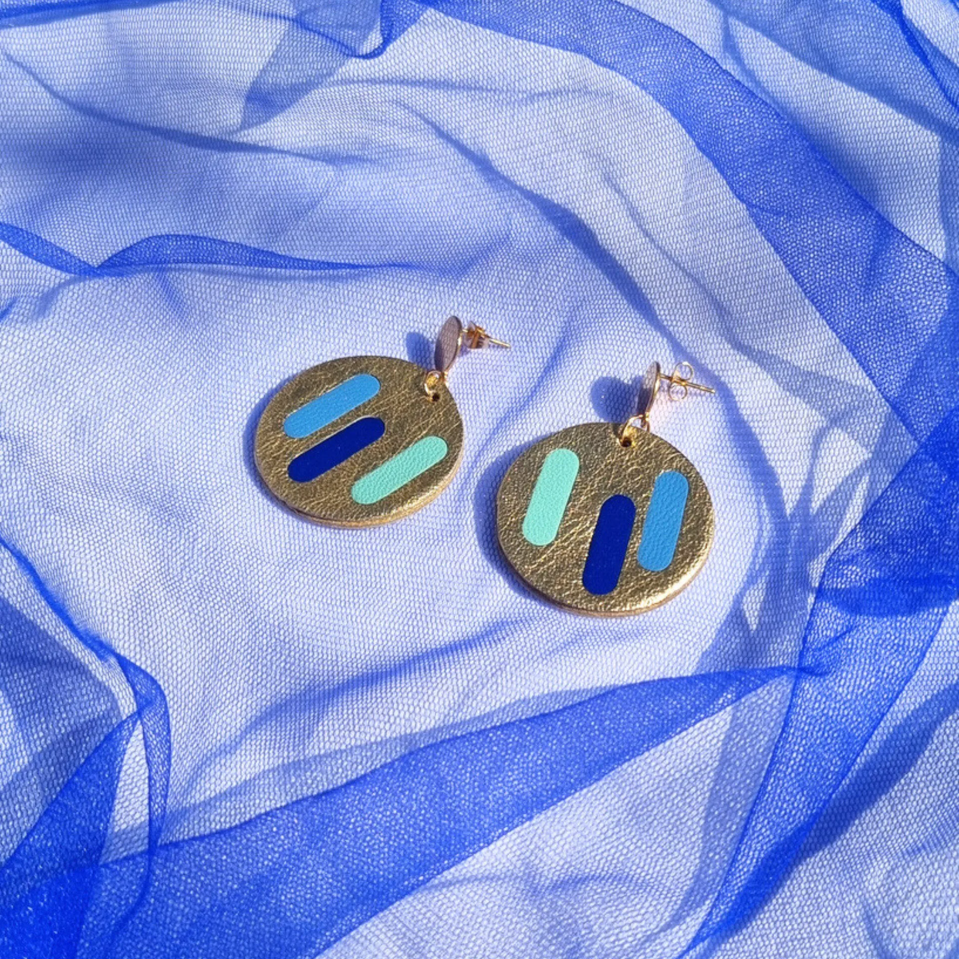 Circular gold leather disc earrings with blue line inlays, displayed on a draped blue mesh fabric. With a gold stud fastening.