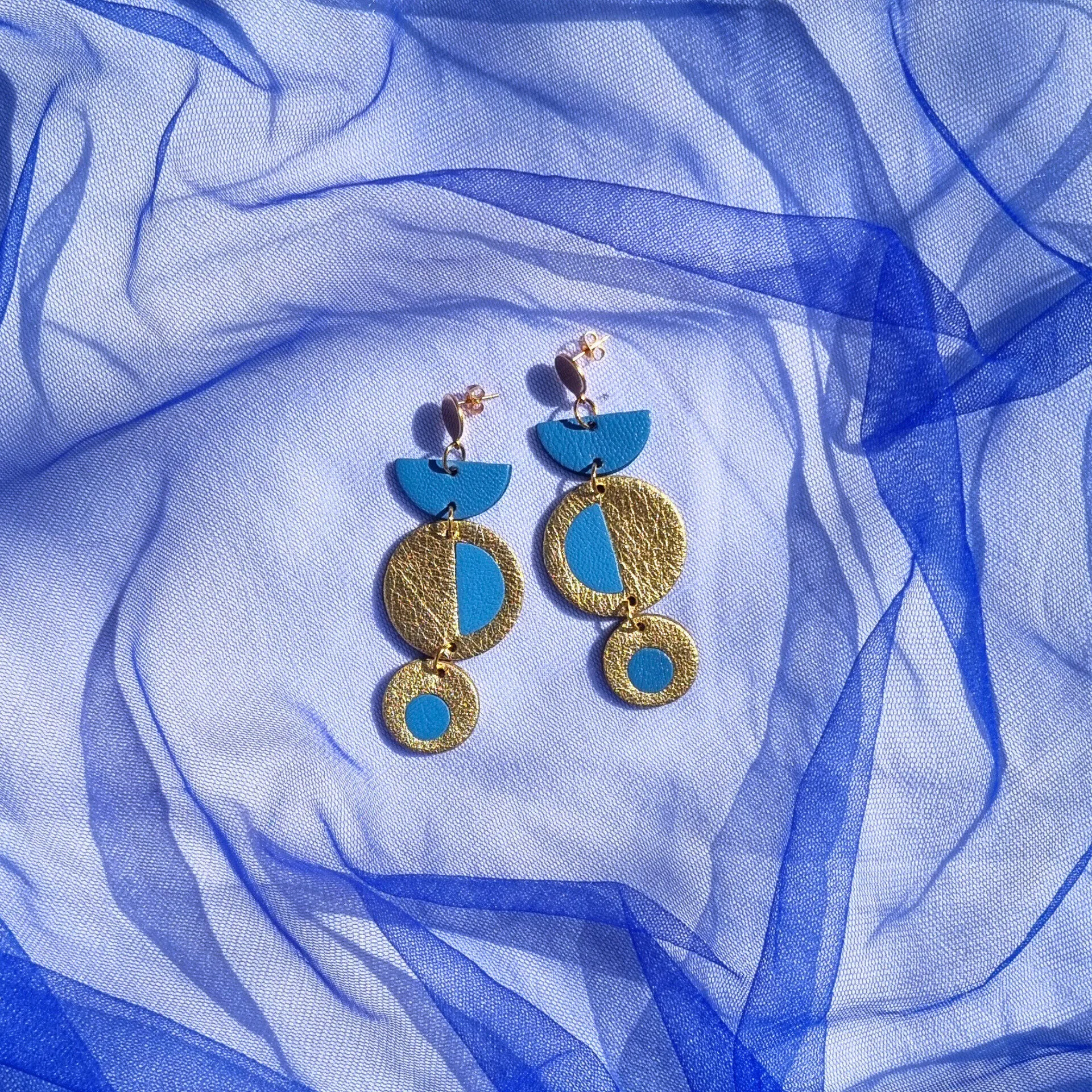 Dangly geometric earrings featuring azure blue and metallic gold leather inlays, displayed on a royal blue mesh fabric background.