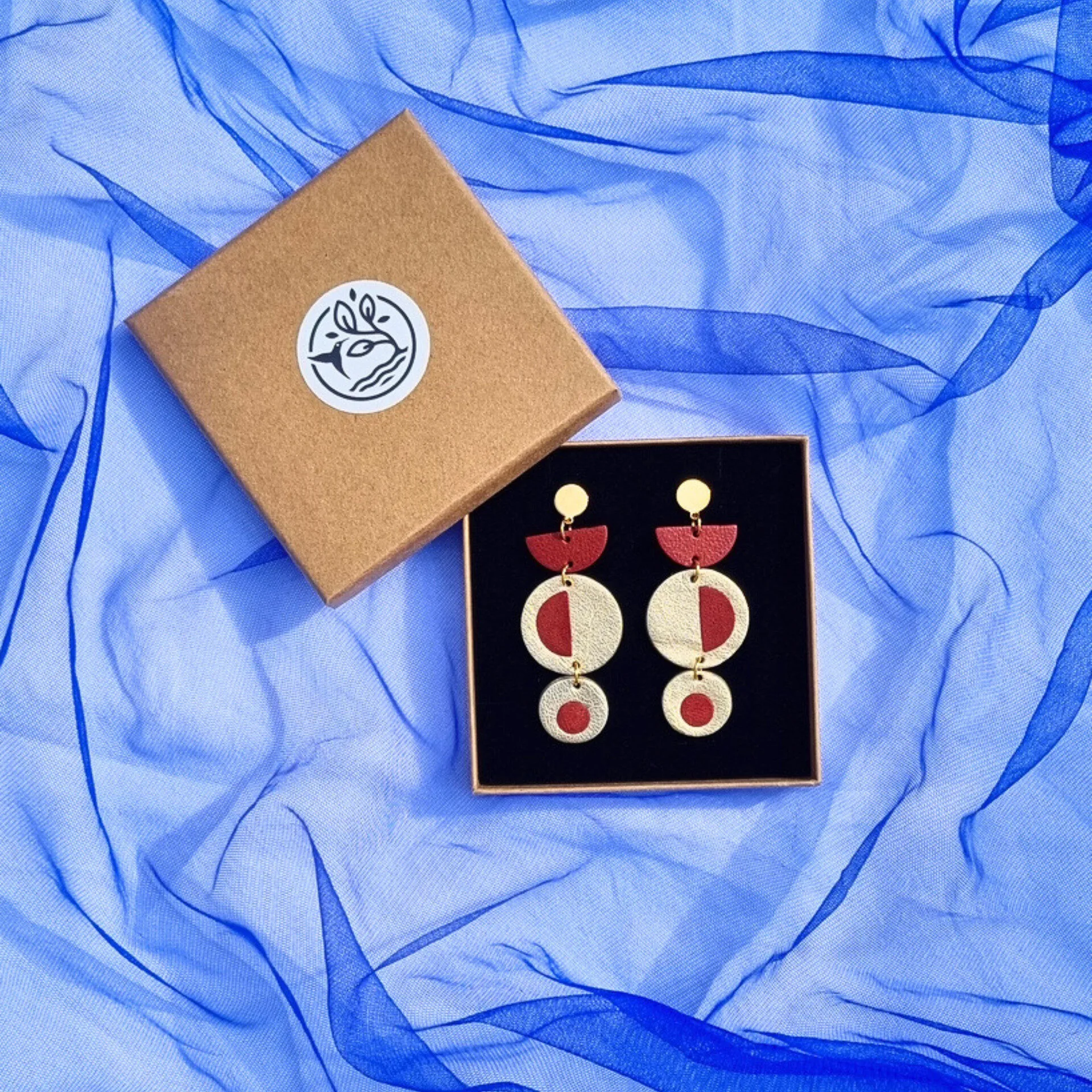Handcrafted geometric dangly earrings with brick red inlayed in metallic gold leather, in a kraft jewellery box on a royal blue mesh fabric background.